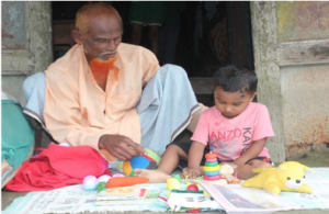 A man with a long red bread sits on the ground this his young child. Both of them are playing with a wide range of toys and learning materials displayed before them on a mat.